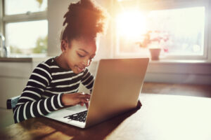 Ethnic girl with a cute afro hairstyle sitting at home using a laptop computer to do her class work and browse the internet, bright sun glow behind